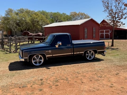 Classic blue and black pickup truck parked at a rural ranch with red barn and wooden fence